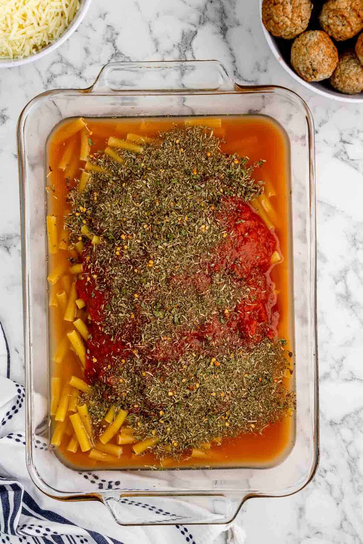 Italian seasoning, chicken stock and pasta sauce poured over uncooked ziti noodles in a baking dish