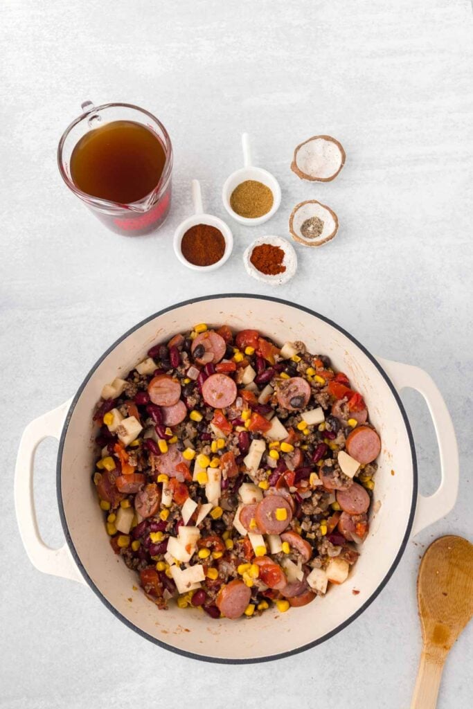 ground beef, sausage slices, beans, corn, tomatoes and potatoes all being stirred together in a pot