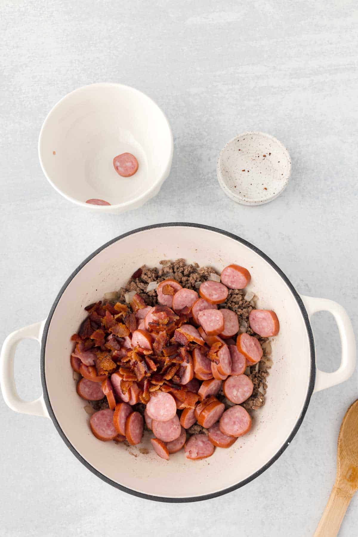 cooked bacon pieces and sausage slices added to a Dutch oven with cooked ground beef