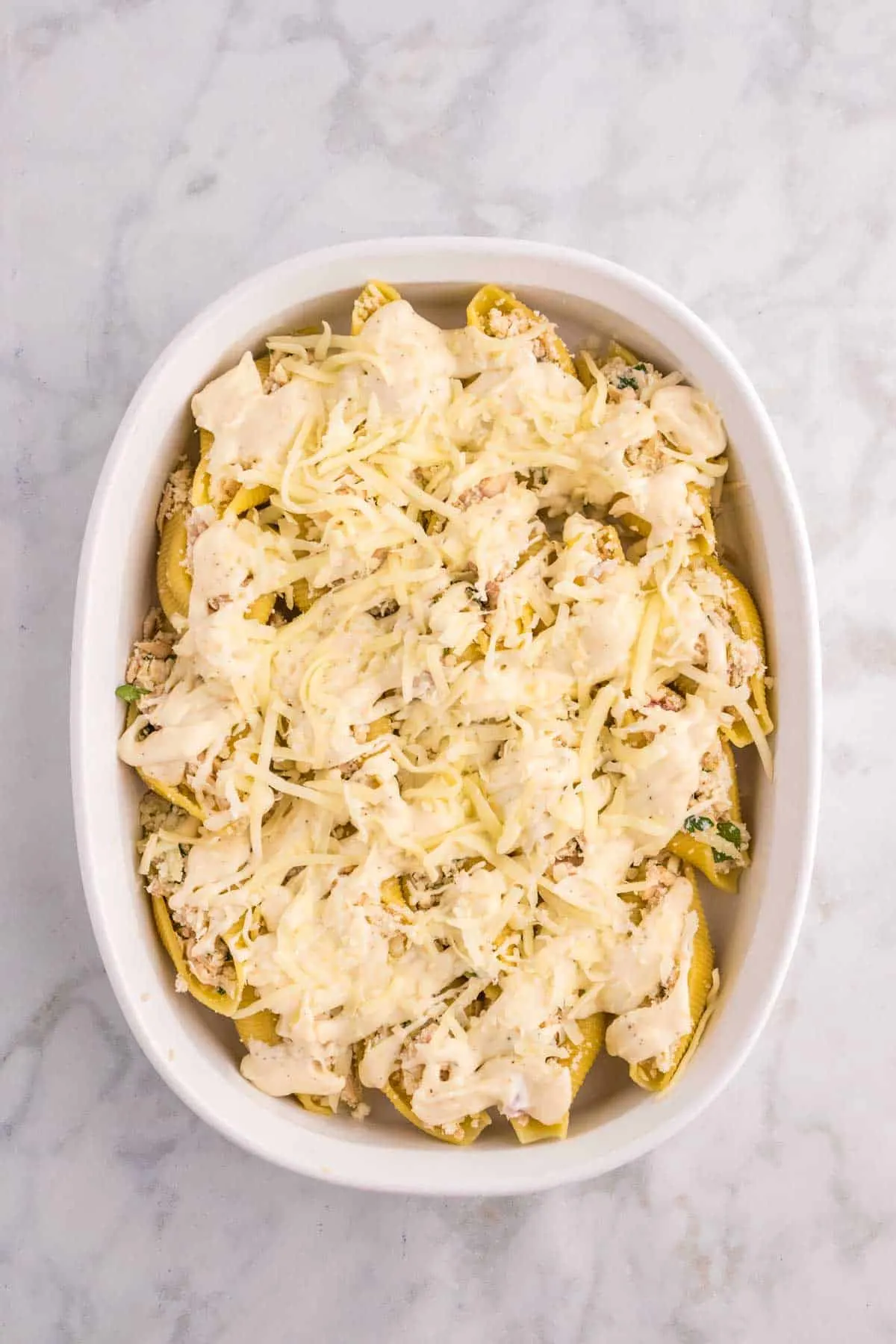 shredded mozzarella and garlic cream sauce on top of tuna stuffed shells in a baking dish