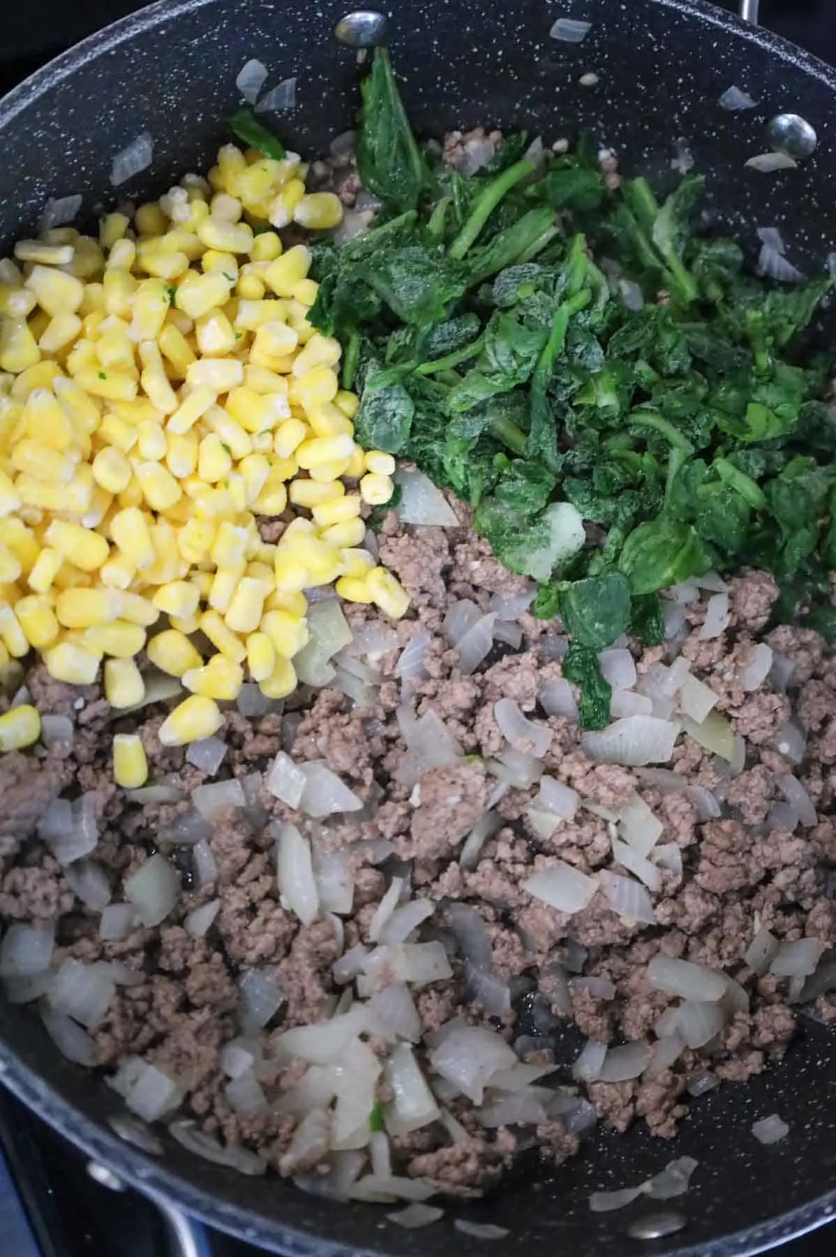 frozen corn and chopped spinach on top of cooked ground beef and diced onions in a skillet
