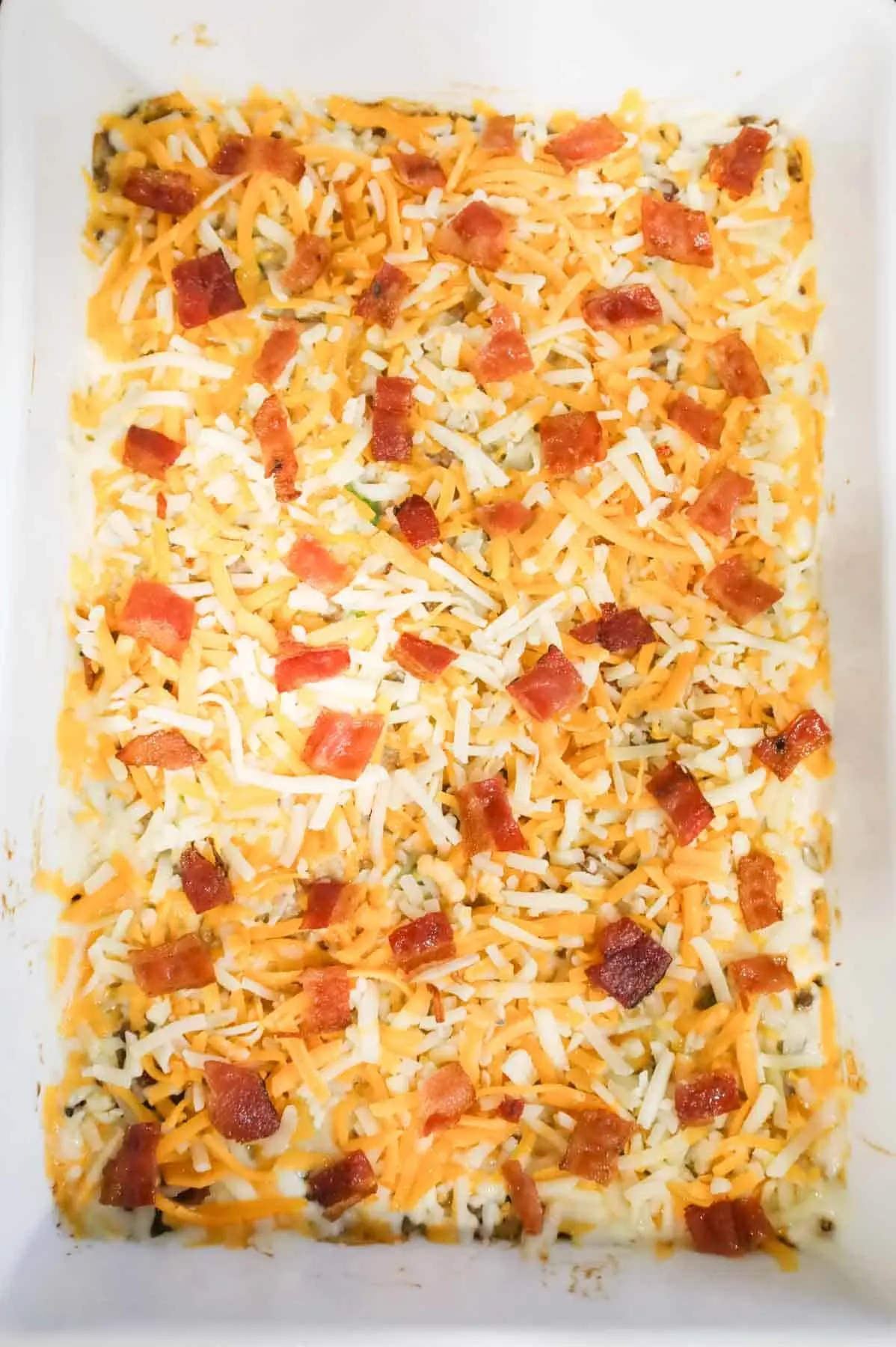 shredded cheese and cooked bacon pieces on top of creamy ground beef and rice mixture in a baking dish