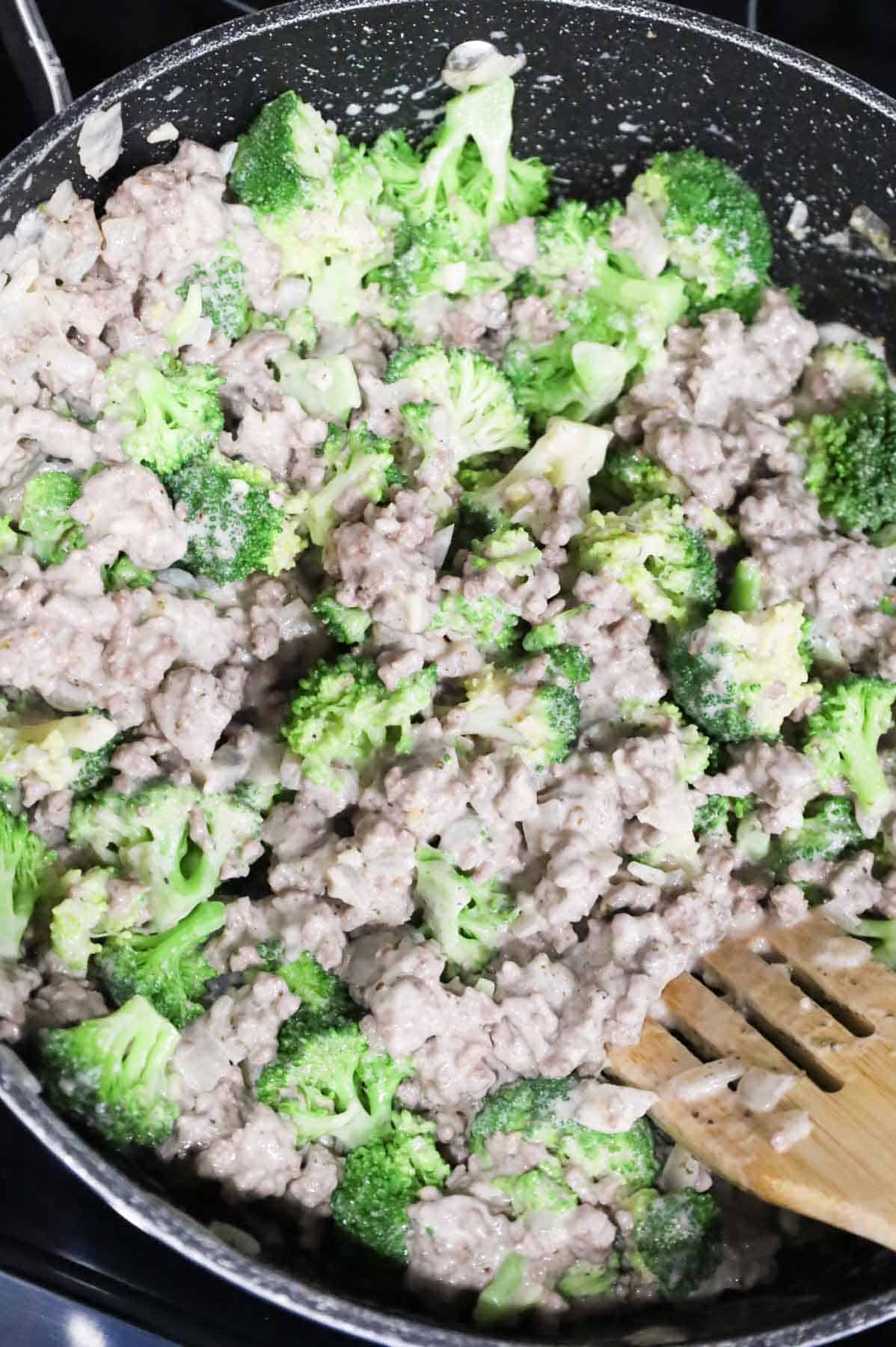broccoli florets being stirred into ground beef and alfredo sauce mixture in a skillet