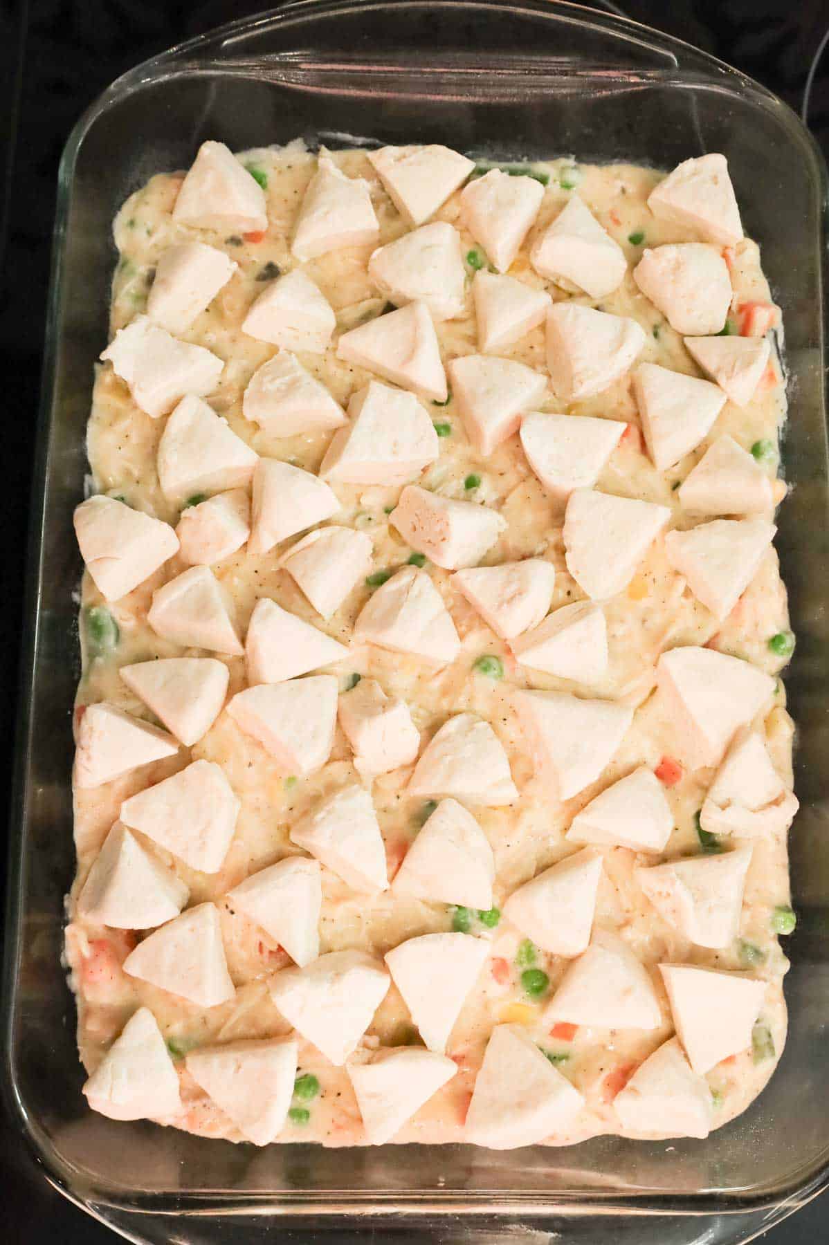 raw Pillsbury biscuit dough pieces on top of creamy chicken and vegetables mixture in a baking dish.