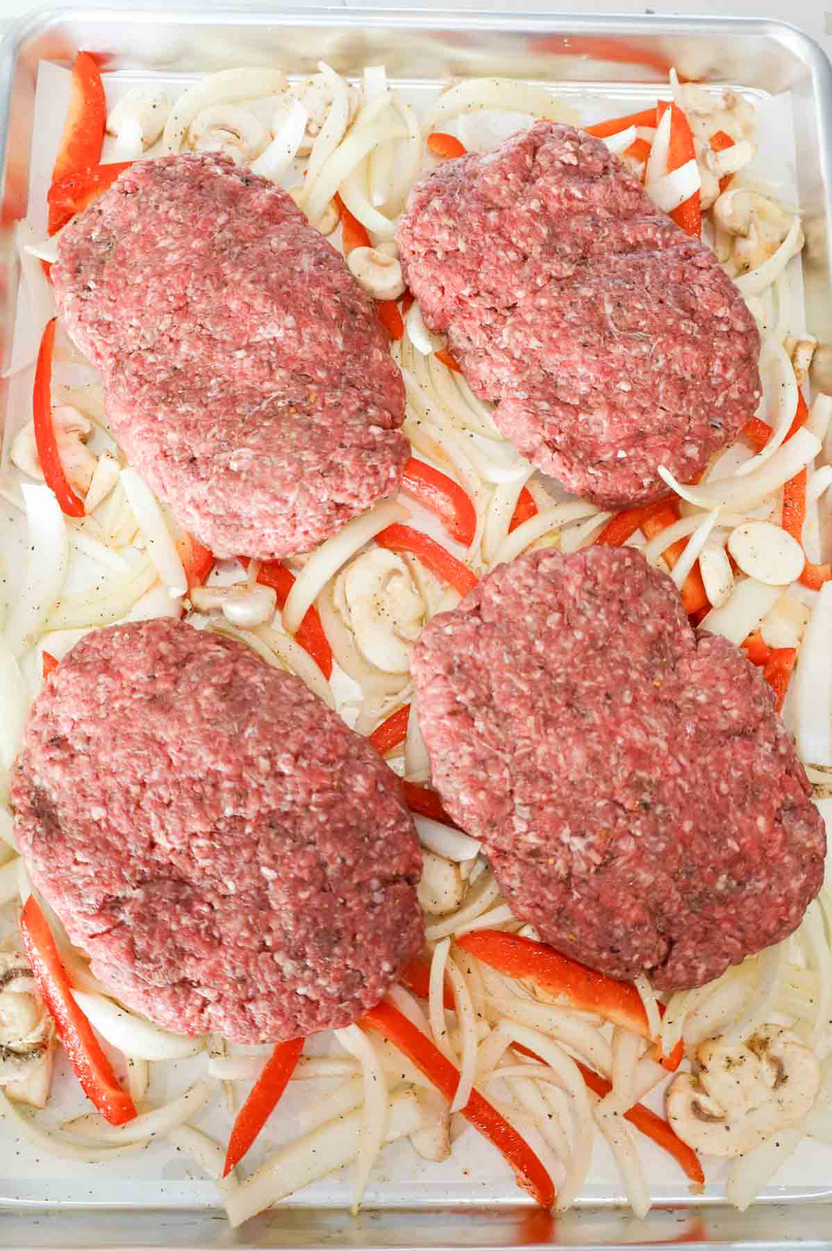 raw ground beef patties on top of sliced onions, red peppers and mushrooms on a baking sheet