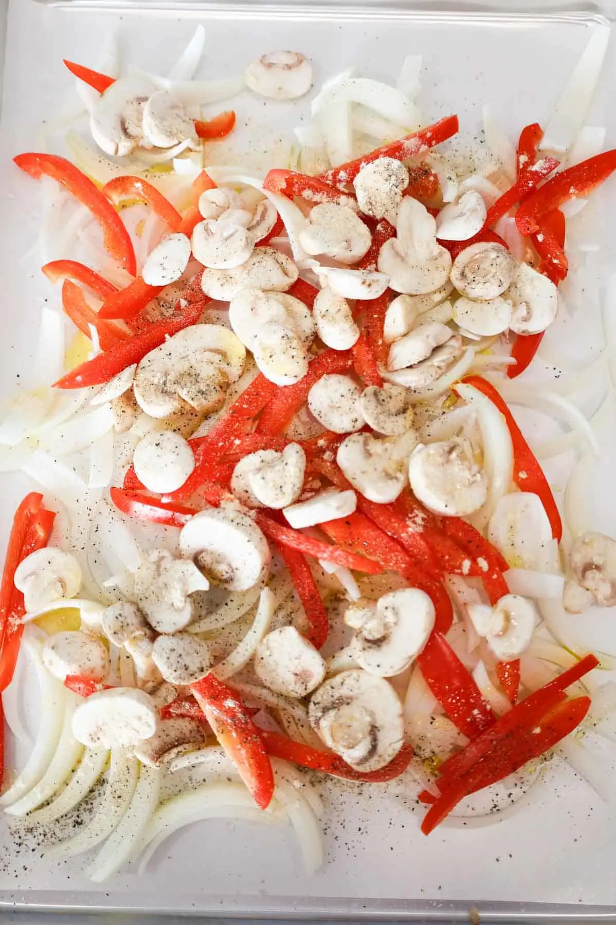 seasoning and olive oil on top of sliced onions, mushrooms and red bell peppers on a parchment lined sheet pan