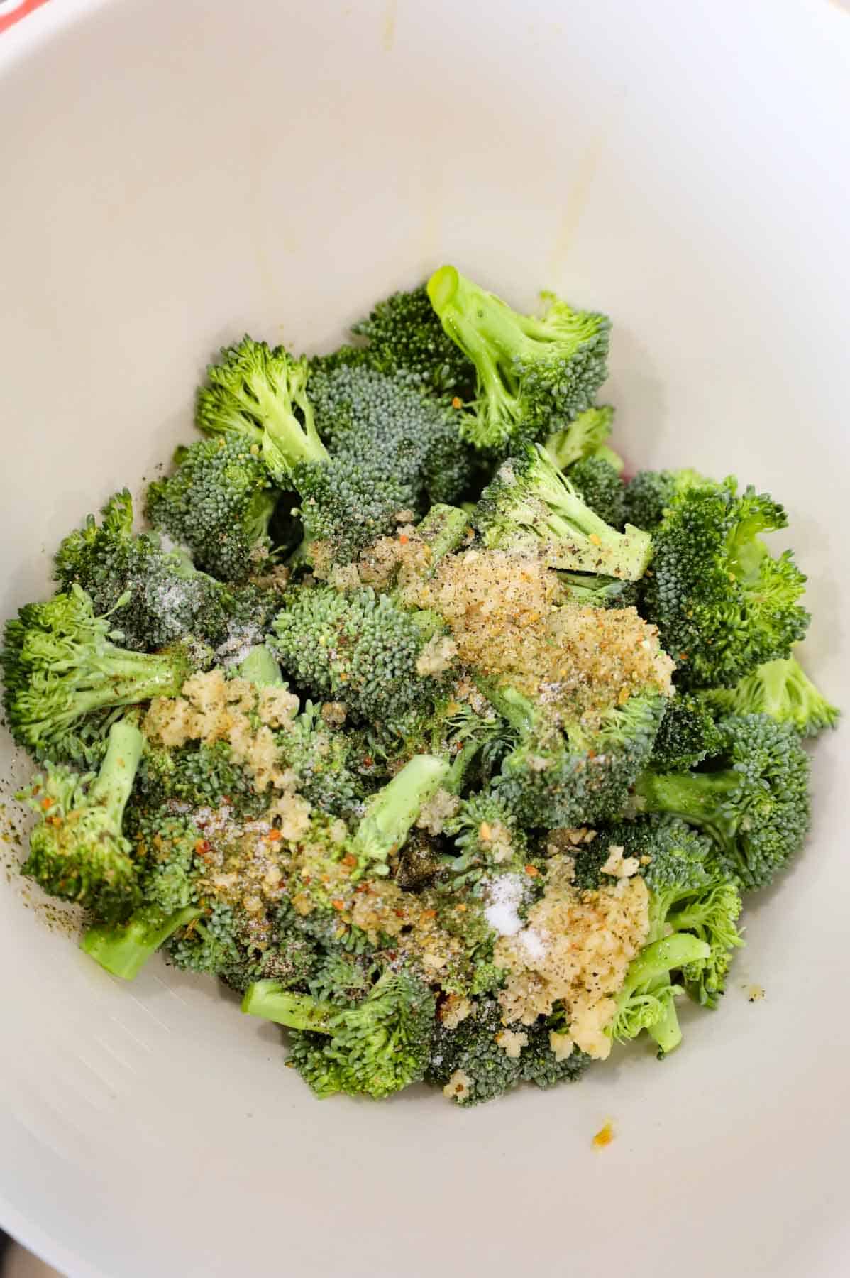 minced garlic, spices and olive oil on top of broccoli florets in a mixing bowl