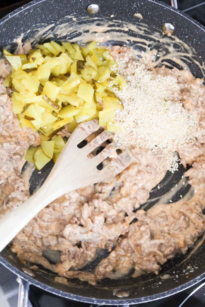 sesame seeds and chopped dill pickles on top of ground beef mixture in a skillet