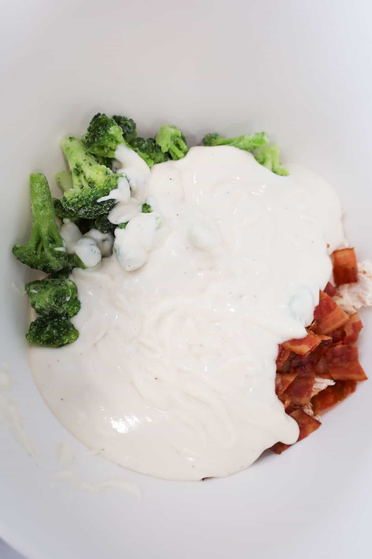alfredo sauce poured over broccoli, chicken and bacon in a mixing bowl