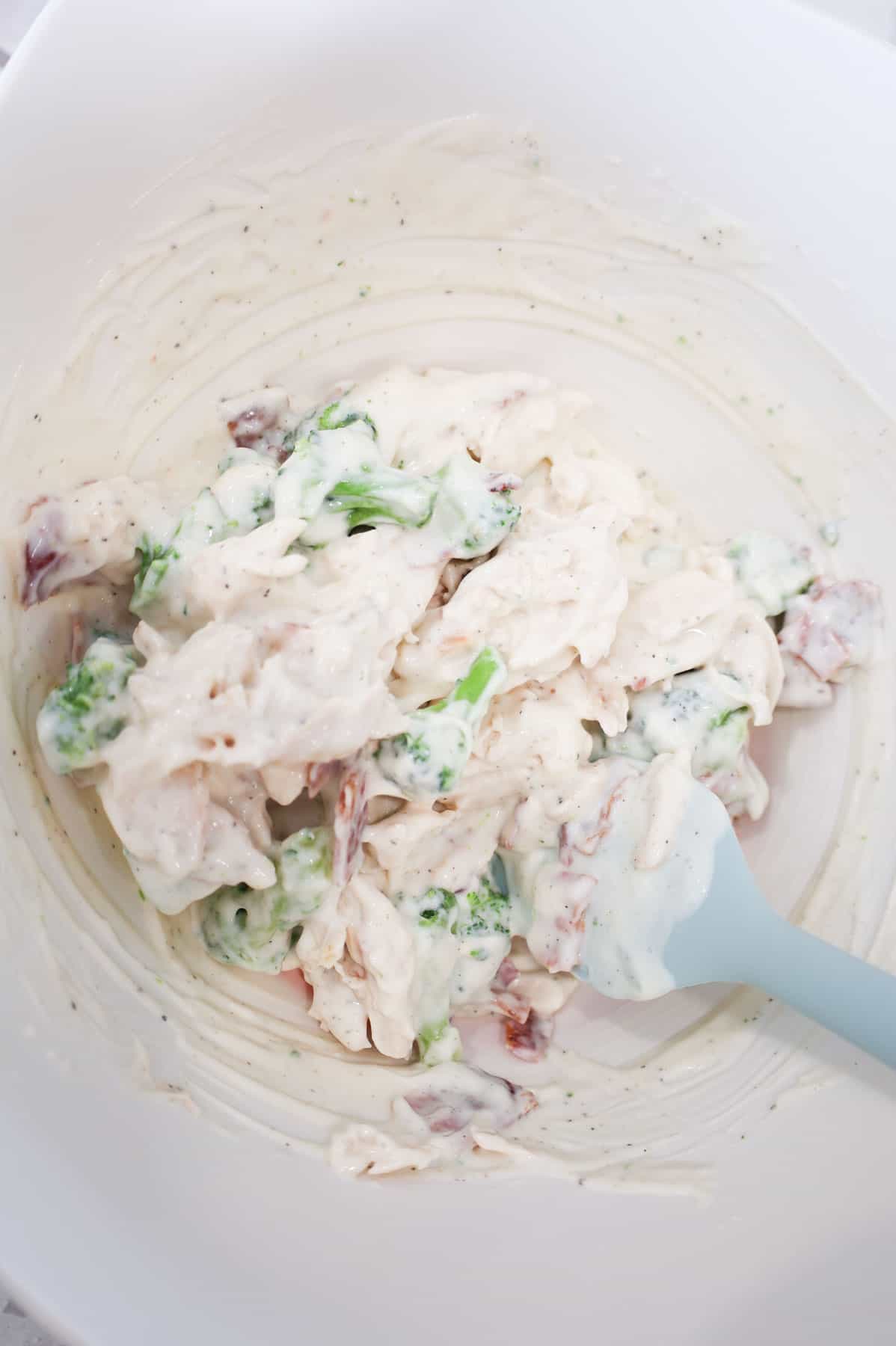 alfredo sauce, bacon, broccoli florets and shredded bacon being stirred together in a mixing bowl