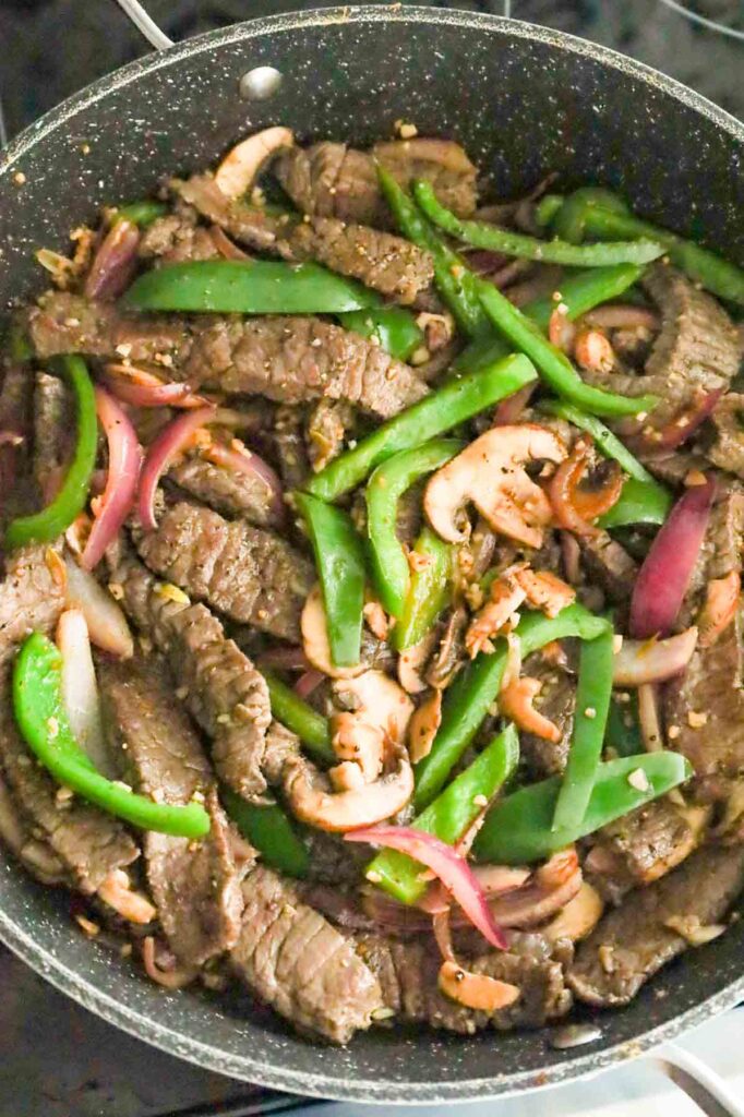steak spice being stirred with steak strips, green peppers, red onions and mushrooms in a skillet