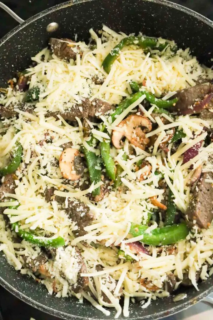 shredded mozzarella and provolone on top of steak slices, green peppers, red onions and mushrooms in a skillet