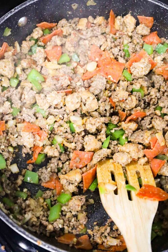 pepperoni being stirred with ground beef, sausage meat and green peppers in a skillet