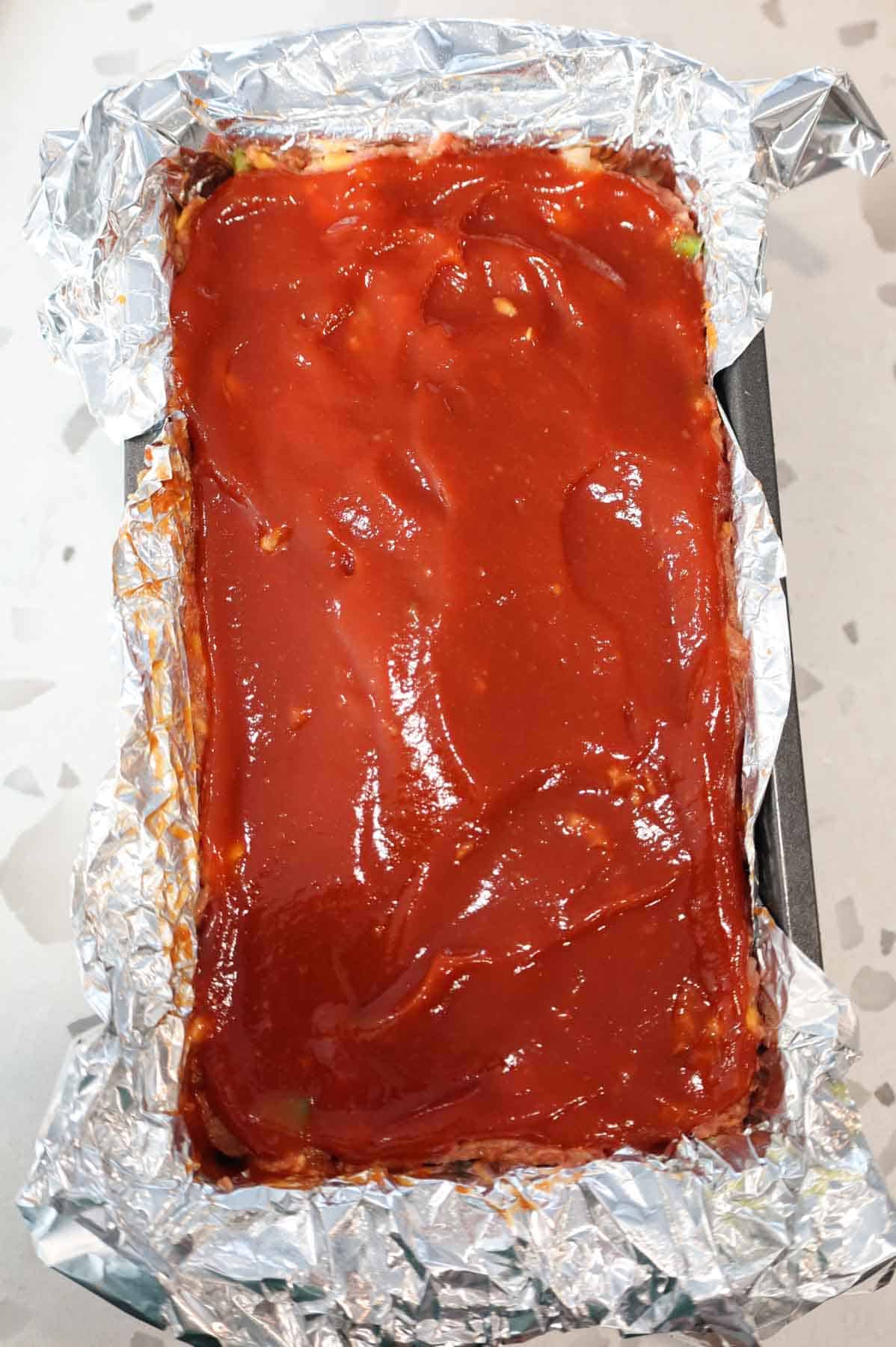 Ketchup glaze spread over top of meatloaf in foil lined loaf pan