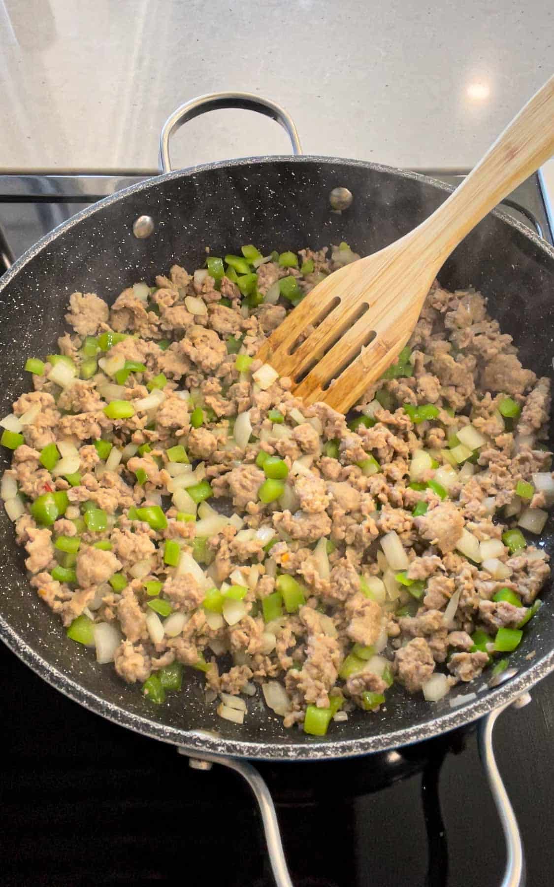 diced green peppers and diced onions being stirred with cooked sausage meat in a skillet