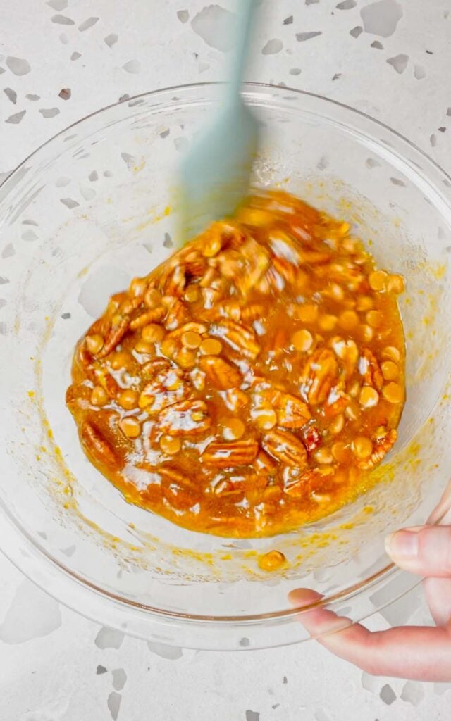 pecan halves and Reese's chips being stirred in to sugary mixture in a mixing bowl