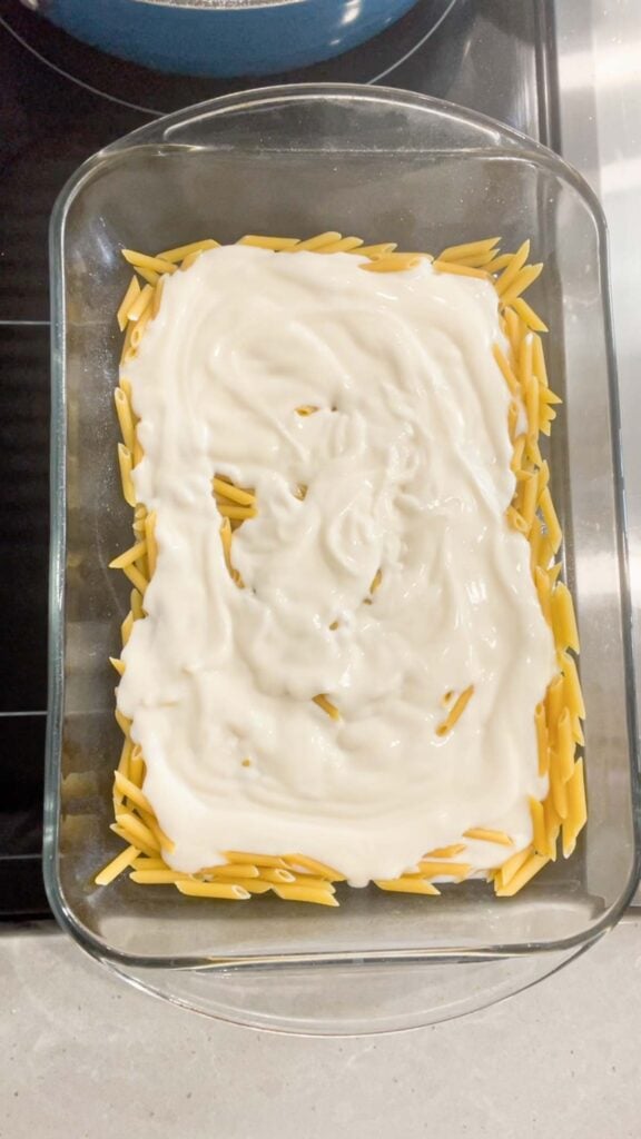 alfredo sauce on top of uncooked mini penne noodles in a baking dish
