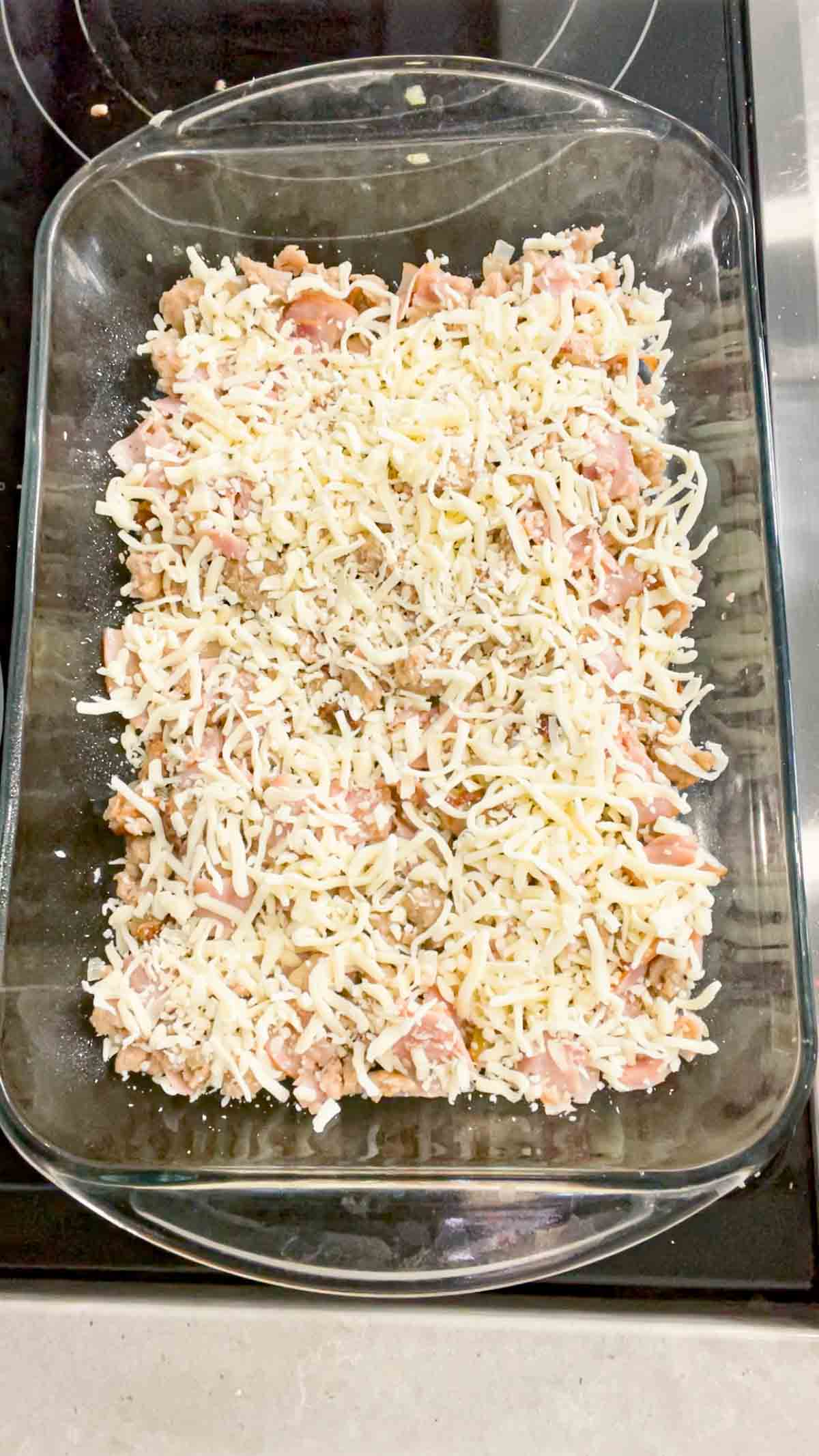 shredded mozzarella cheese on top of cooked sausage and ham mixture in a baking dish
