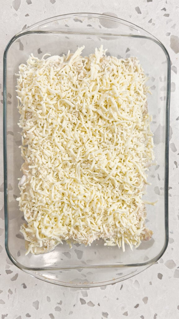 shredded mozzarella cheese on top of chicken and frozen pierogies in a glass baking dish