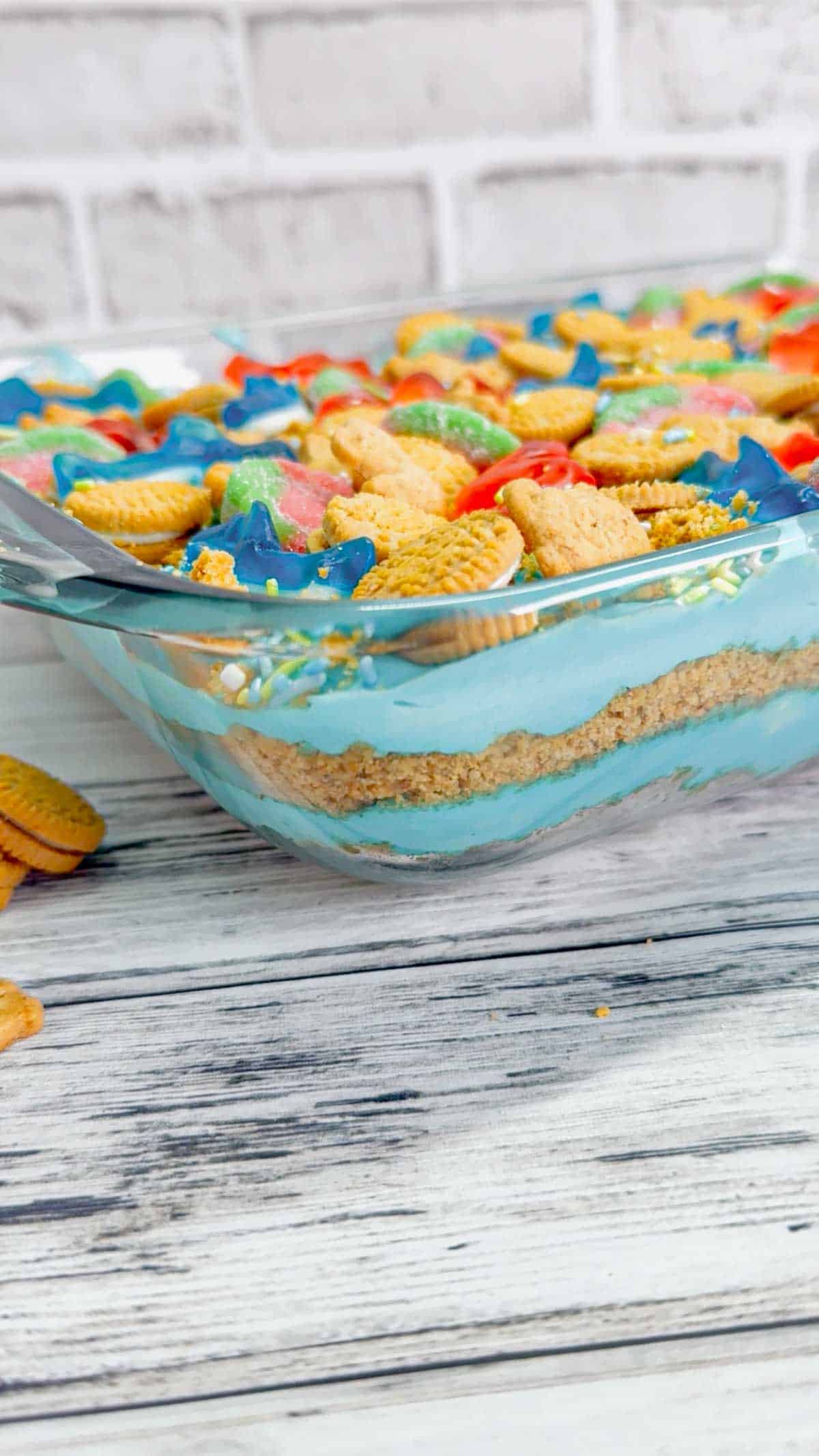 Summer Dirt Cake is an easy no bake dessert recipe perfect for parties and potlucks. This beach themed dirt cake is made with golden Oreo and graham cracker crumbs, layered with a delicious cream cheese and vanilla pudding mixture and all topped with mini cookies and beach themed gummy candies.