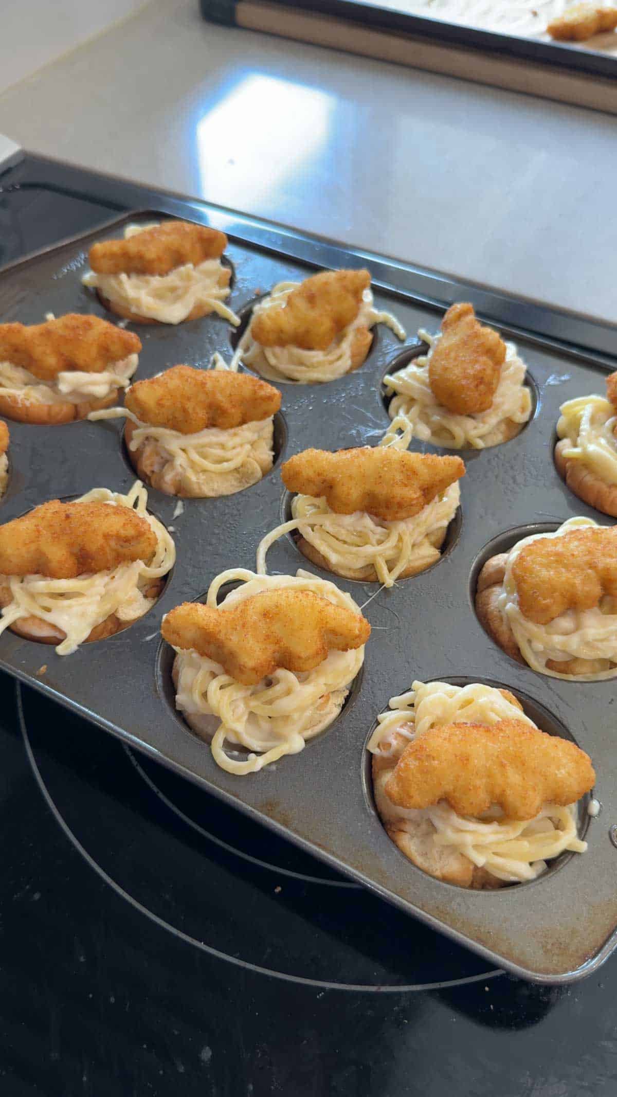 dino nuggets on top of alfredo spaghetti stuffed into hamburger cups in a muffin tin