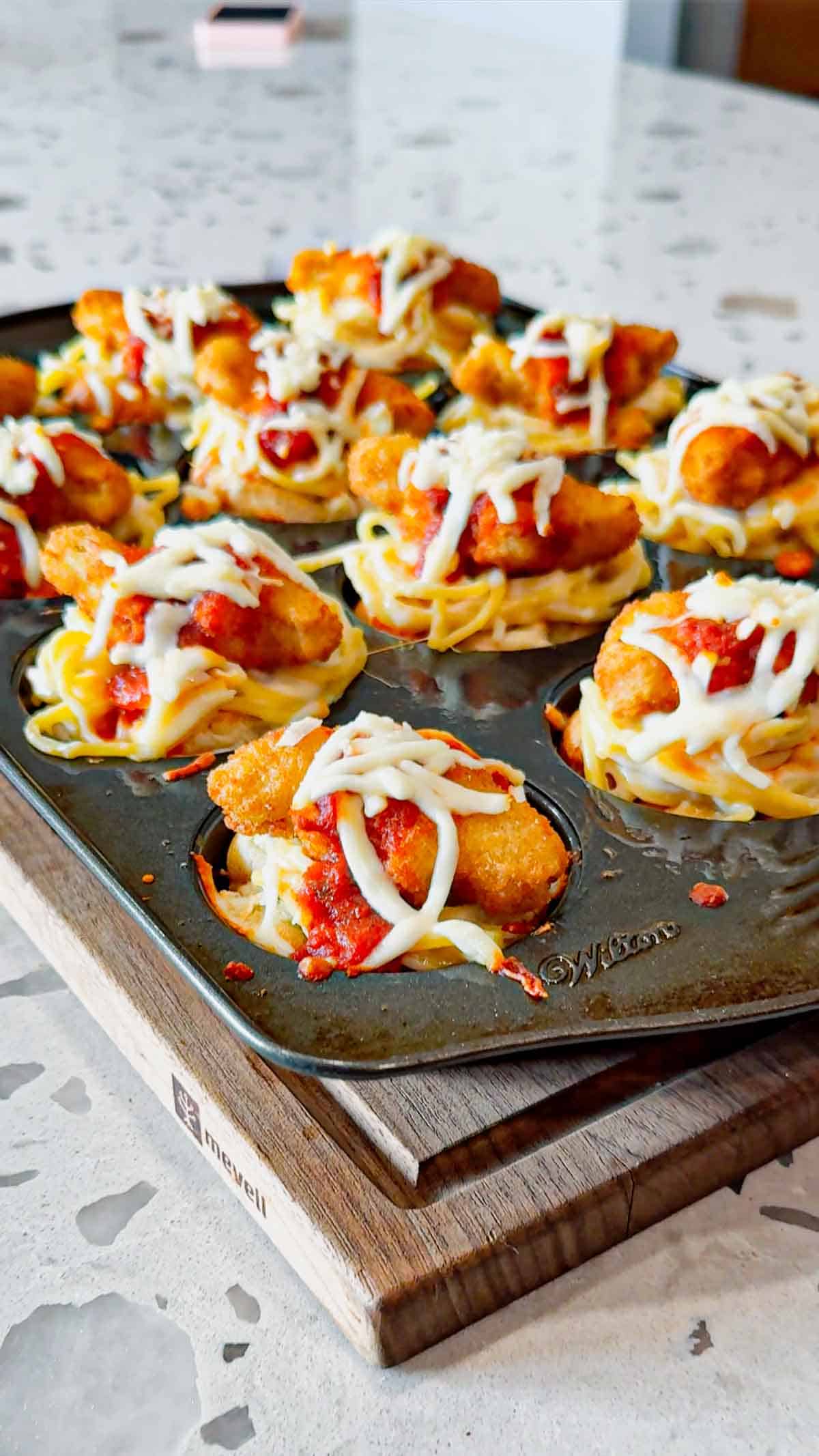 Dino Nugget Chicken Parm Cups are a fun and easy muffin tin dinner the whole family will love. Hamburger buns are spread with garlic butter, baked in muffin tins and stuffed with creamy alfredo spaghetti and topped with dino nuggets, marinara and mozzarella.