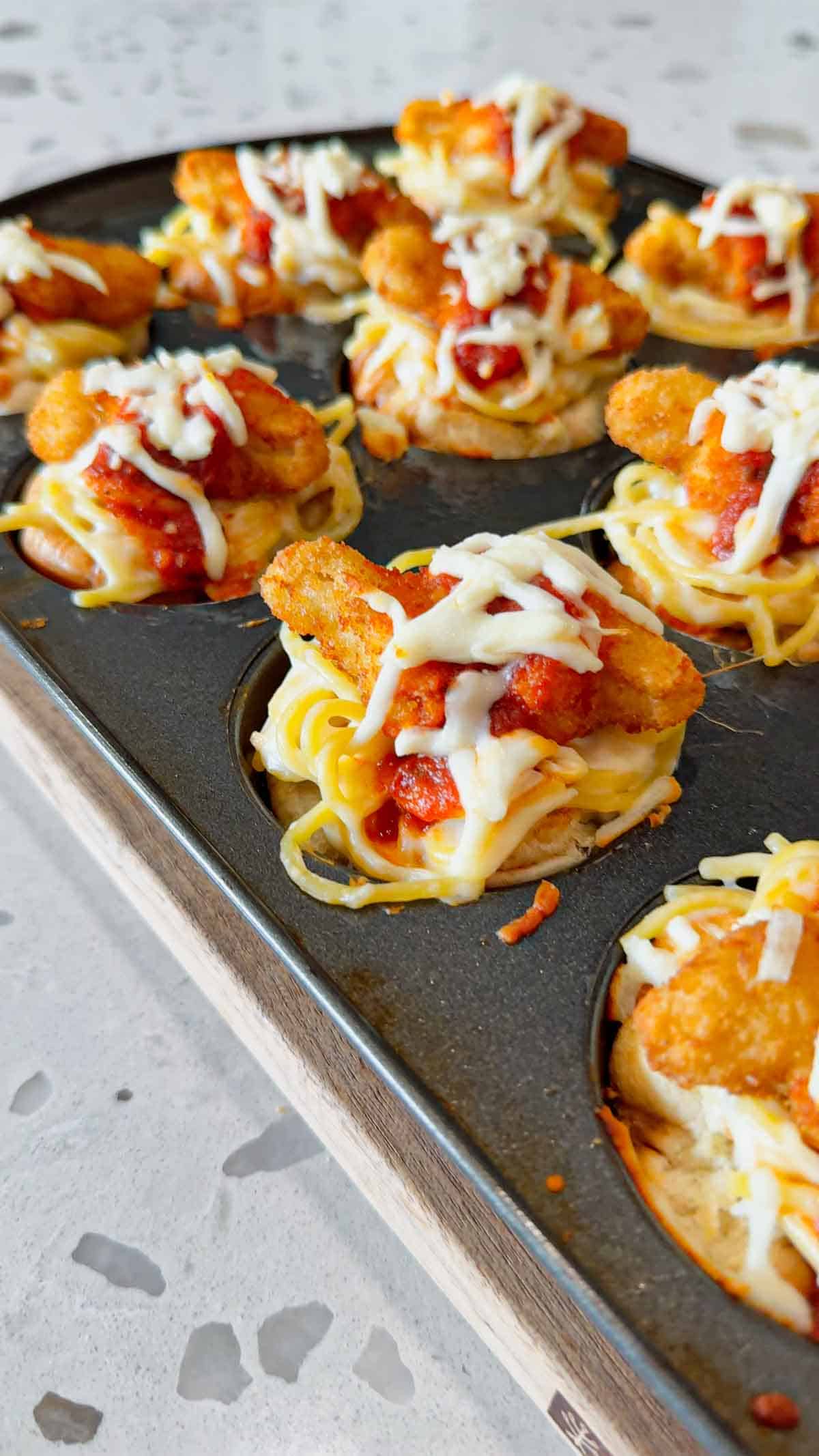 Dino Nugget Chicken Parm Cups are a fun and easy muffin tin dinner the whole family will love. Hamburger buns are spread with garlic butter, baked in muffin tins and stuffed with creamy alfredo spaghetti and topped with dino nuggets, marinara and mozzarella.