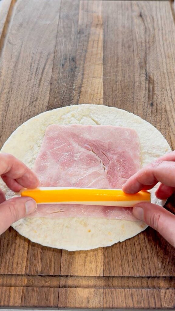 sliced ham and a stick of string cheese on a flour tortilla on a cutting board