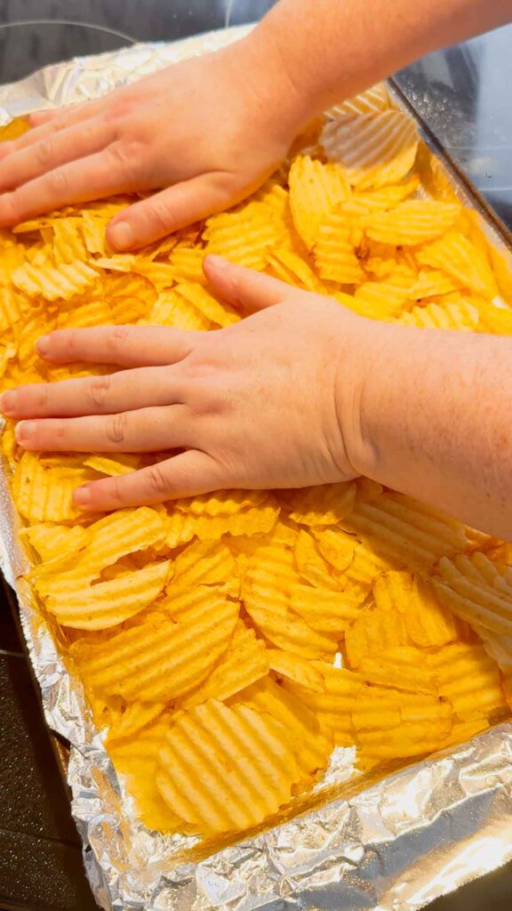 hands pressing down on ruffled potato chips on a foil lined baking sheet