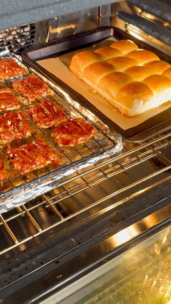 mini mcrib patties coated in bbq sauce in oven next to a tray of hawaiian rolls