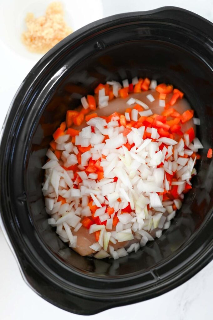 diced onions and red bell peppers on top of raw chicken breasts in a slow cooker