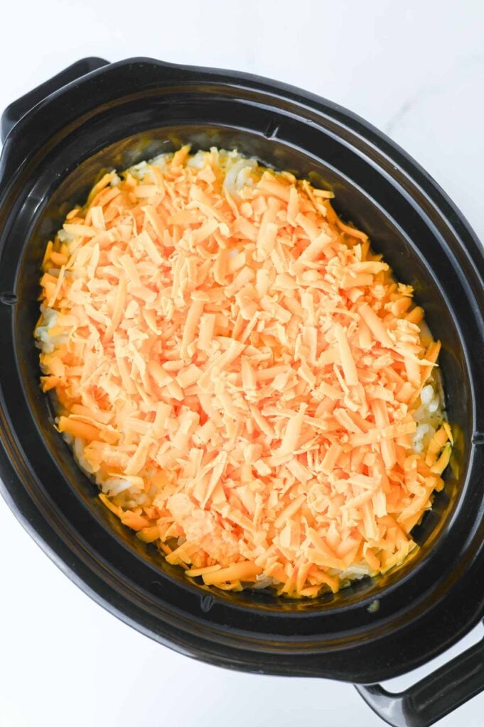 shredded cheddar cheese on top of hashbrown casserole in a slow cooker