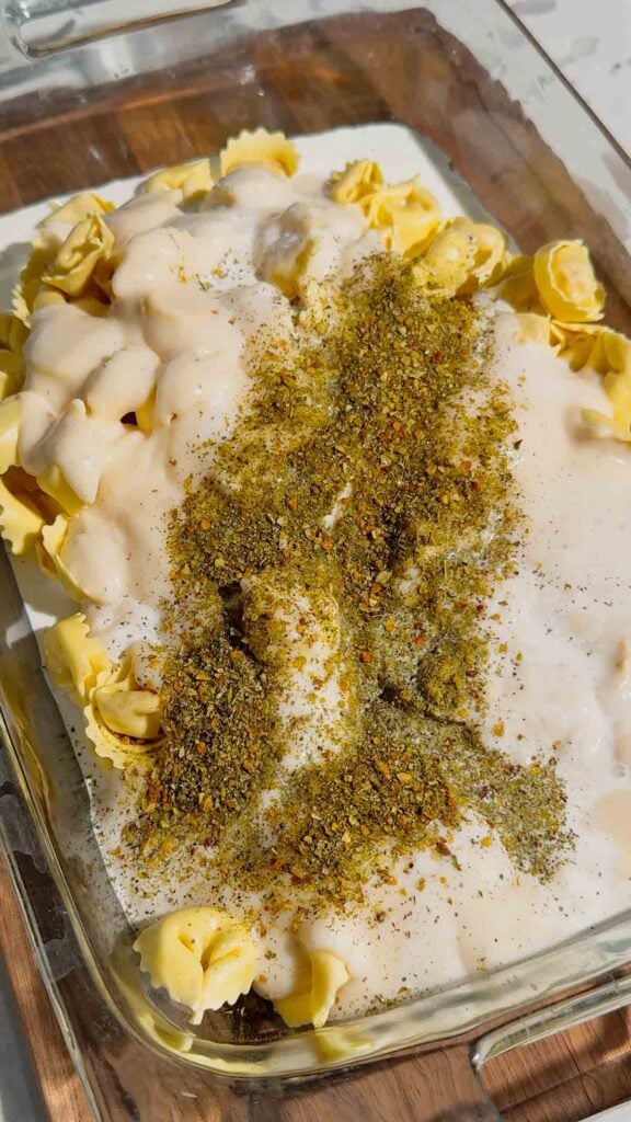 italian seasoning and alfredo sauce on top of tortellini in a baking dish