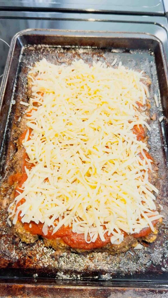 shredded mozzarella cheese on top of pizza sauce and meatloaf on a bkaing sheet