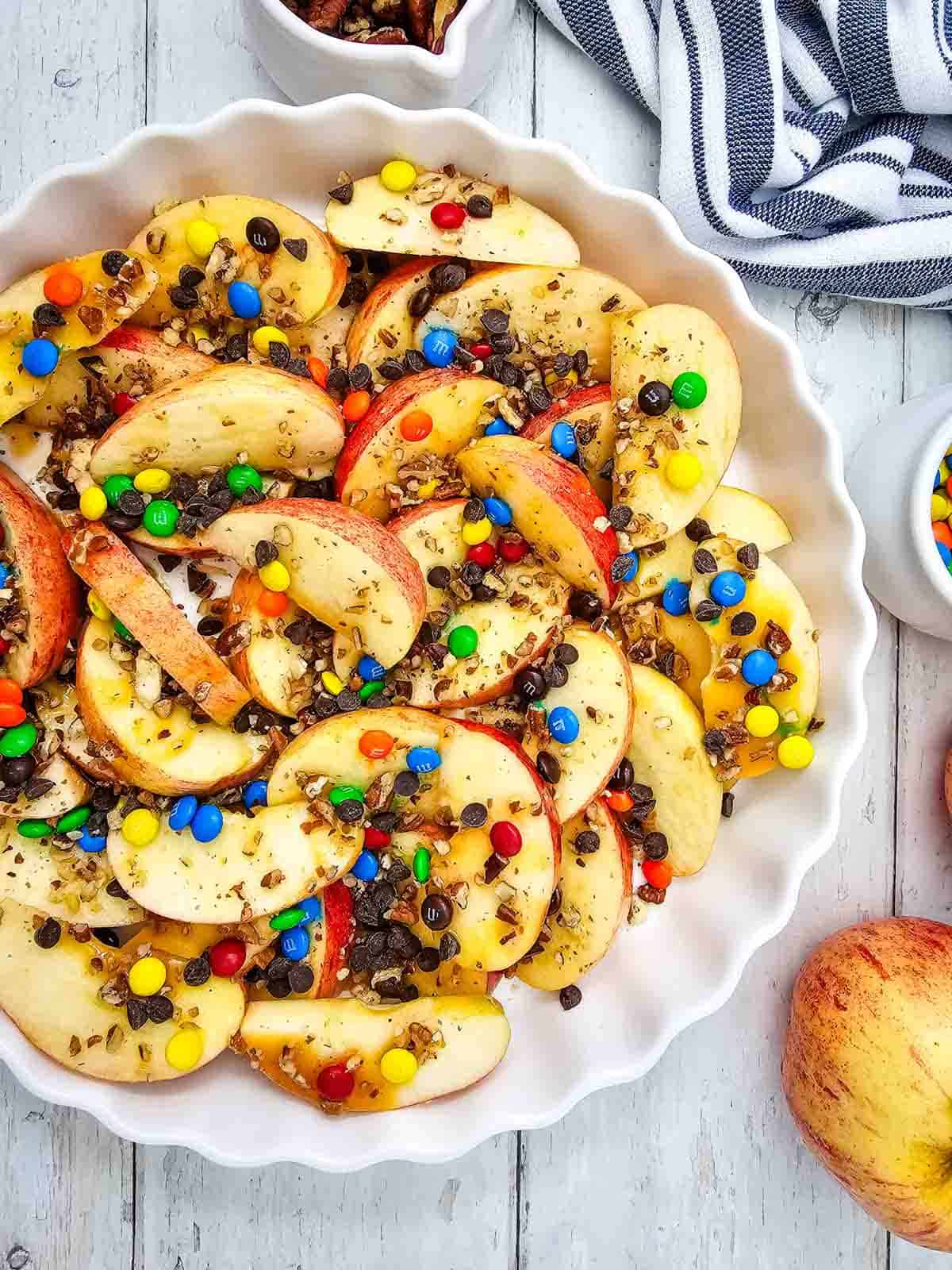 Looking for a fun, easy, and crowd-pleasing snack? These Apple Nachos are the ultimate no-bake dessert recipe made with crisp fresh apples, rich caramel sauce, mini chocolate chips, colorful mini M&Ms, and crunchy pecans. Ready in just 10 minutes, they're perfect for easy fall desserts, healthy-ish snacks for kids, after school snacks, party food ideas, apple dessert recipes, sweet snack recipes, Halloween treats, Thanksgiving snacks, and quick family desserts. Whether you're searching for caramel apple recipes, apple slices with toppings, no bake apple desserts, or a simple fruit dessert everyone will love, these loaded Apple Nachos are a delicious way to enjoy fresh apples with a fun candy twist. Save this easy recipe for your next snack craving or celebration!