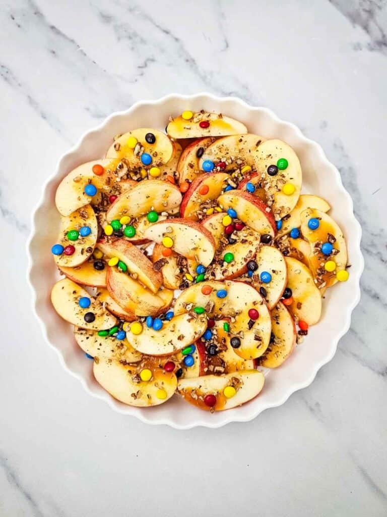 Looking for a fun, easy, and crowd-pleasing snack? These Apple Nachos are the ultimate no-bake dessert recipe made with crisp fresh apples, rich caramel sauce, mini chocolate chips, colorful mini M&Ms, and crunchy pecans. Ready in just 10 minutes, they're perfect for easy fall desserts, healthy-ish snacks for kids, after school snacks, party food ideas, apple dessert recipes, sweet snack recipes, Halloween treats, Thanksgiving snacks, and quick family desserts. Whether you're searching for caramel apple recipes, apple slices with toppings, no bake apple desserts, or a simple fruit dessert everyone will love, these loaded Apple Nachos are a delicious way to enjoy fresh apples with a fun candy twist. Save this easy recipe for your next snack craving or celebration!