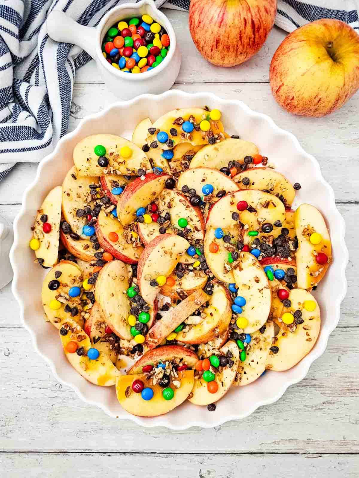 Looking for a fun, easy, and crowd-pleasing snack? These Apple Nachos are the ultimate no-bake dessert recipe made with crisp fresh apples, rich caramel sauce, mini chocolate chips, colorful mini M&Ms, and crunchy pecans. Ready in just 10 minutes, they're perfect for easy fall desserts, healthy-ish snacks for kids, after school snacks, party food ideas, apple dessert recipes, sweet snack recipes, Halloween treats, Thanksgiving snacks, and quick family desserts. Whether you're searching for caramel apple recipes, apple slices with toppings, no bake apple desserts, or a simple fruit dessert everyone will love, these loaded Apple Nachos are a delicious way to enjoy fresh apples with a fun candy twist. Save this easy recipe for your next snack craving or celebration!
