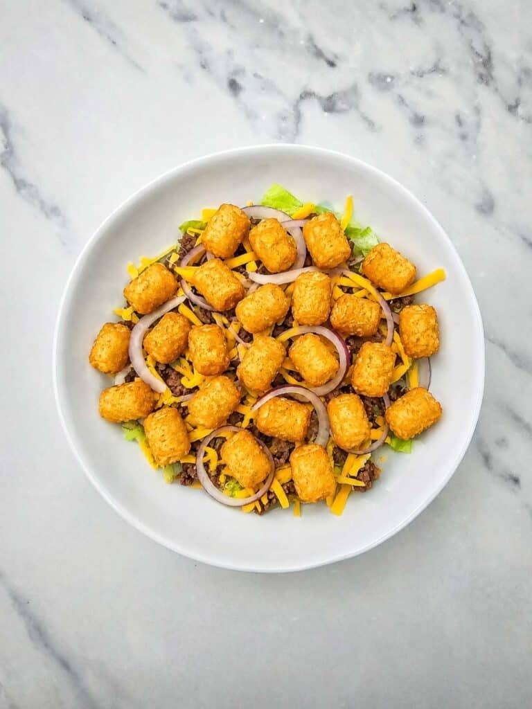 cooked tater tots on top of onions, cheddar, ground beef and lettuce in a bowl