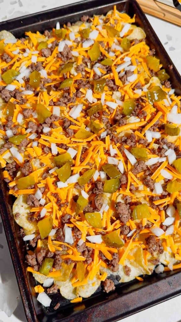 chopped pickles, onions, cheddar cheese and cooked ground beef on top of kettle chips on a baking sheet
