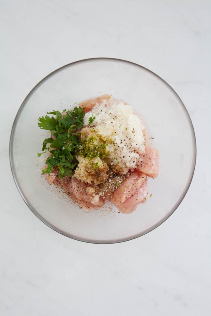onion, cumin, lime zest, cilantro and garlic on top of raw chicken breast chunks in a glass bowl