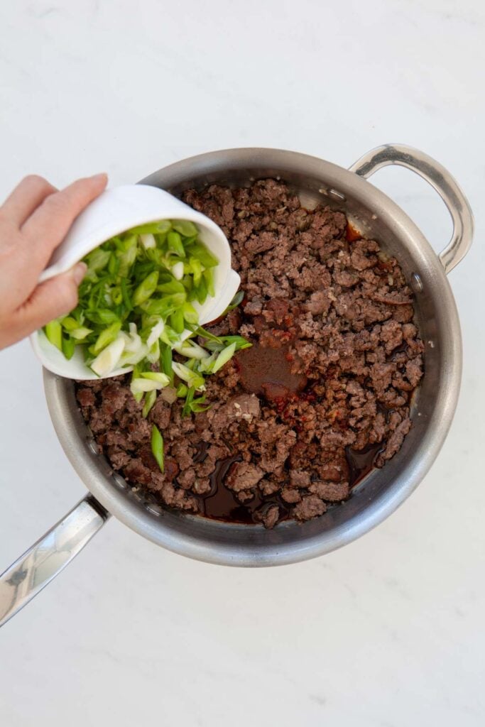 pouring chopped green onions into skillet with cooked ground beef and soy sauce mixture