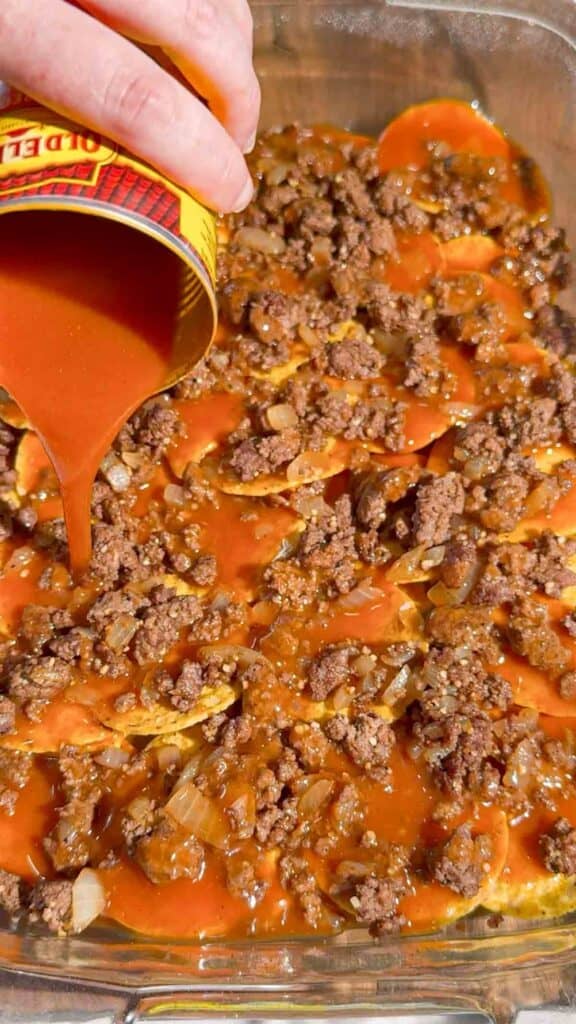 pouring a can of red enchilada sauce over ground beef and tortilla chips in a baking dish