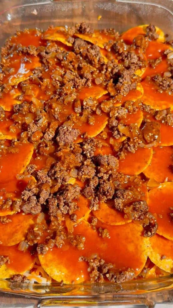 red enchilada sauce poured over ground beef and tortilla chips in a baking dish
