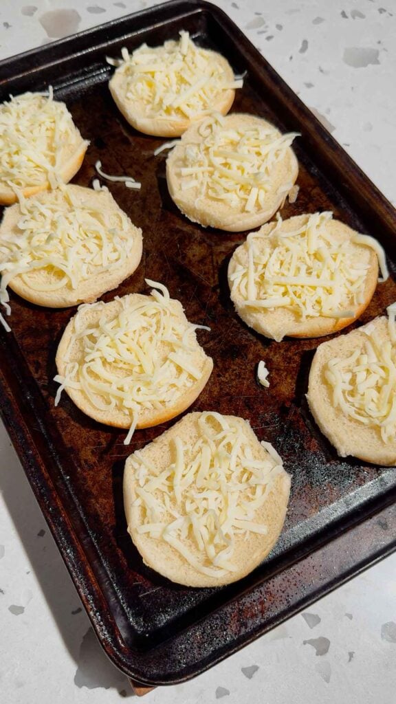 shredded mozzarella cheese on top of burger buns on a baking sheet