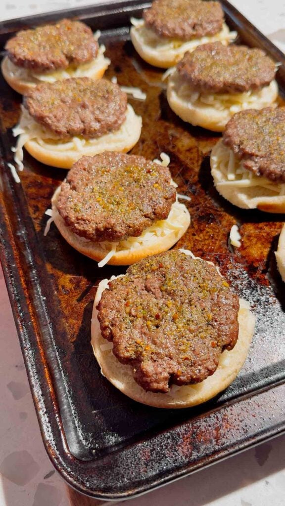 hamburger patties on top of cheese and burger buns on a baking sheet