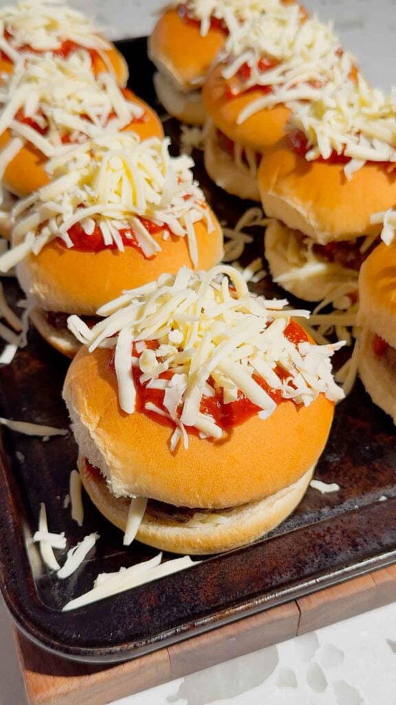 shredded mozzarella and pizza sauce on top of burgers on a baking sheet