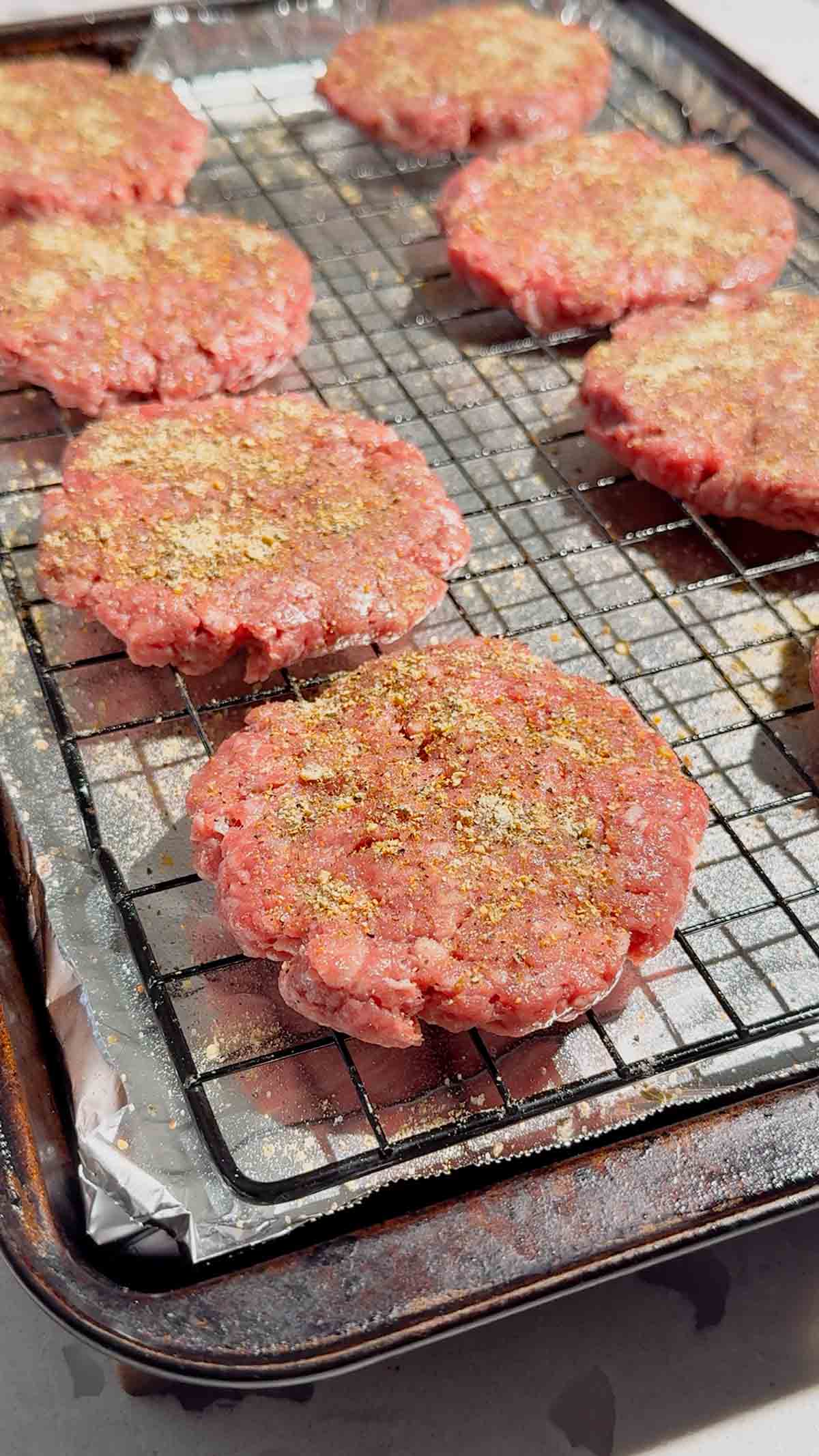 raw seasoned hamburger patties on a rack on a foil lined baking sheet