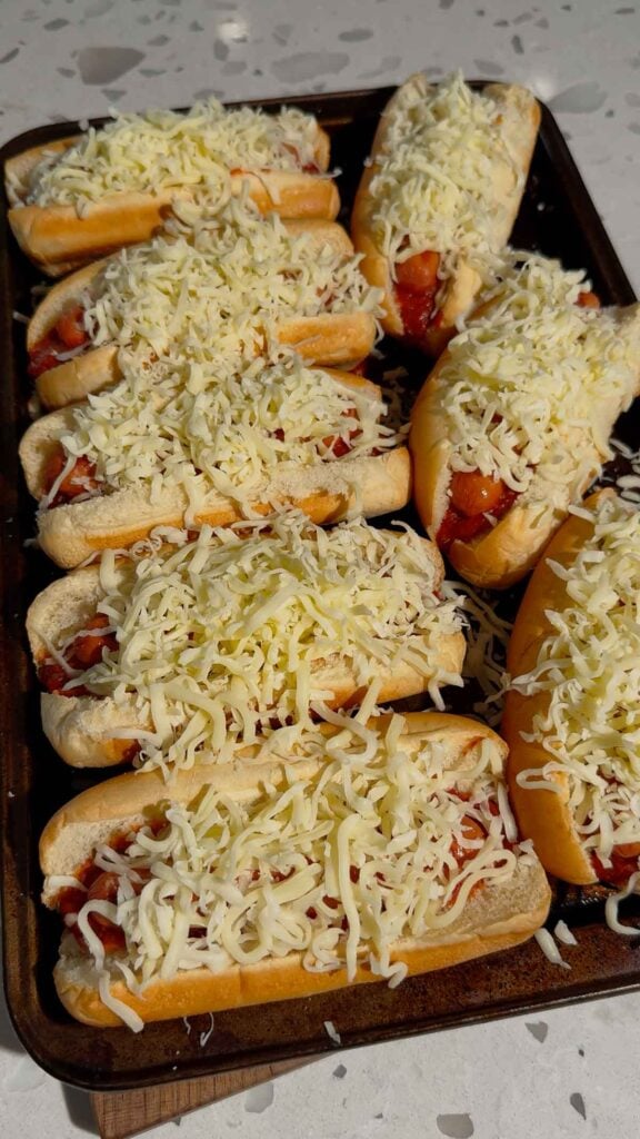 shredded mozzarella on top of hot dogs on a baking sheet