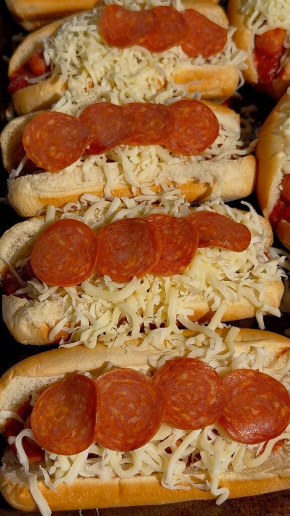 pepperoni and shredded mozzarella on top of hot dogs on a baking sheet