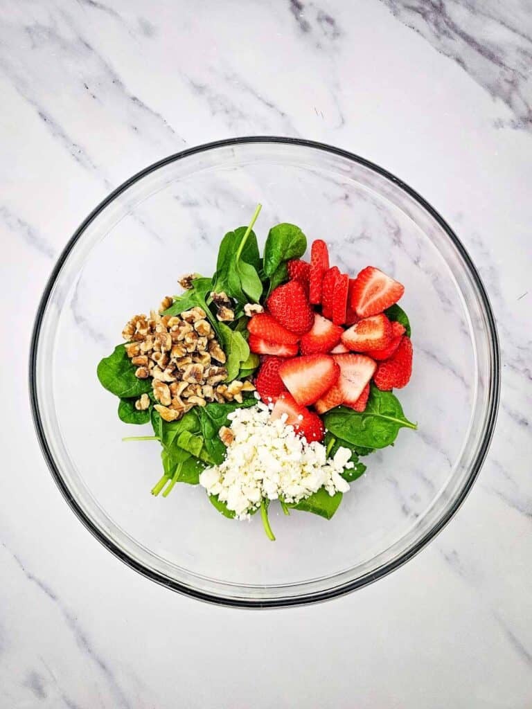 crumbled feta, walnut pieces, sliced strawberries and spinach in a glass mixing bowl
